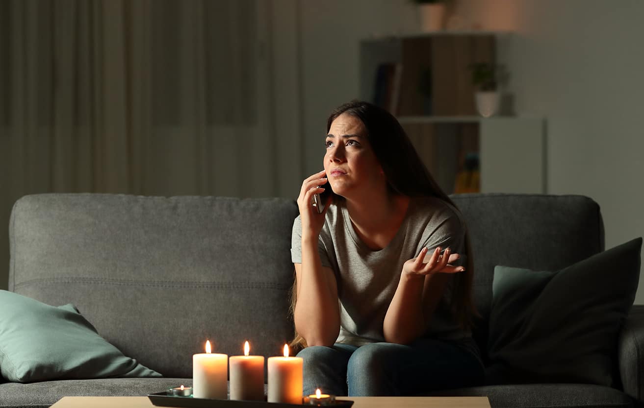 Woman sitting on a sofa, talking on the phone with candles lit during a power outage, highlighting energy solutions by Castaways Energy.