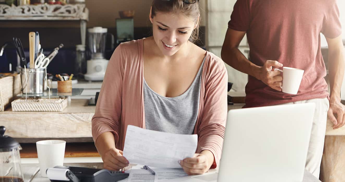 Smiling woman reviewing documents at a kitchen table, with a man holding a coffee cup standing nearby, highlighting energy savings from Castaways Energy.