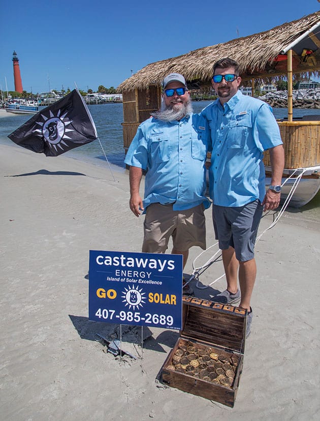 Photo of two Castaways team members standing in front on the beach with Castaways sign and treasure chest.